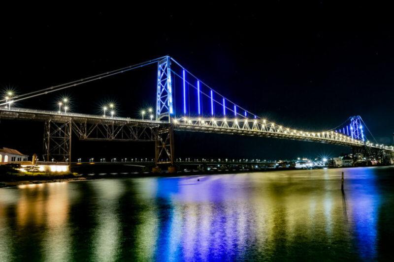 Fotografia noturna da Ponte Hercílio Luz, em Florianópolis, iluminada por luzes azuis e brancas. A estrutura metálica se reflete na água abaixo, criando um efeito de cores vibrantes, principalmente em tons de azul, amarelo e verde. À esquerda, uma pequena construção iluminada compõe a margem. O céu está escuro e sem nuvens, com algumas luzes ao fundo, representando a cidade.