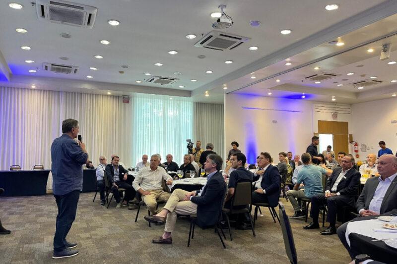 Fotografia de um evento corporativo em uma sala de conferências iluminada. Em primeiro plano, à esquerda, um homem de costas segura um microfone enquanto fala ao público. O público é composto por cerca de vinte homens, muitos vestidos formalmente, sentados em mesas redondas com toalhas brancas e taças. Ao fundo, há cortinas brancas e paredes com iluminação azul. Alguns participantes observam atentamente, enquanto outros conversam entre si. Equipamentos de áudio e vídeo, como uma câmera no tripé, são visíveis ao fundo.