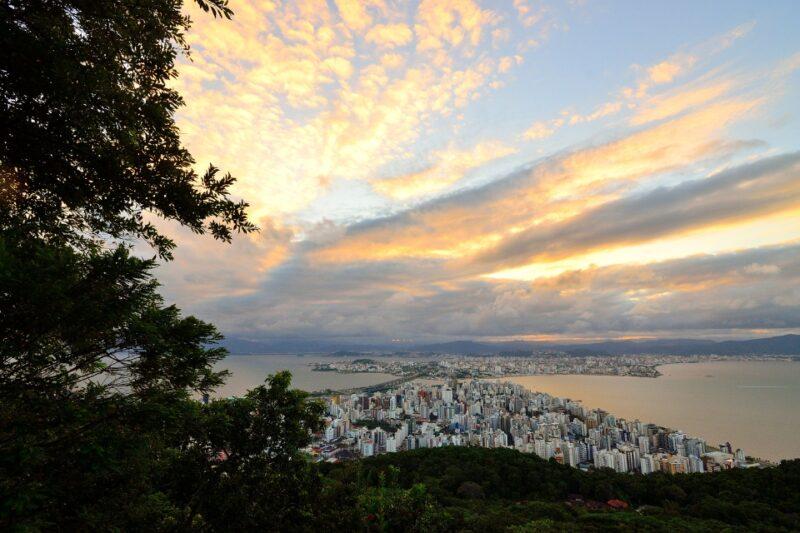 Fotografia de uma paisagem urbana vista do alto, mostrando uma cidade à beira-mar cercada por vegetação e montanhas ao fundo. No primeiro plano, há árvores que emolduram parcialmente a cena. Abaixo, destaca-se um conjunto de edifícios altos e compactos, refletindo uma área urbana densa. À direita, o mar tranquilo reflete a luz do céu, enquanto o horizonte é delineado por montanhas distantes. O céu está repleto de nuvens iluminadas pelo pôr do sol, em tons de laranja, amarelo e azul claro, criando uma atmosfera vibrante e serena.