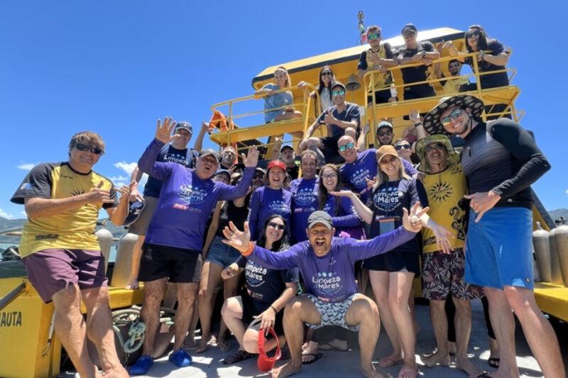 Fotografia de um grupo de aproximadamente **vinte pessoas** em uma embarcação amarela durante o dia, sob um céu azul claro. Elas estão sorrindo, fazendo poses e olhando para a câmera. Muitos vestem camisetas roxas com os dizeres "Limpeza dos Mares", indicando uma ação de conscientização ou limpeza ambiental. Alguns usam chapéus, óculos escuros e roupas leves apropriadas para um ambiente ensolarado. Ao fundo, parte da embarcação é visível, com mais pessoas posicionadas em uma área superior do barco, segurando na grade. O grupo demonstra alegria e espírito de união, com poses descontraídas, como braços erguidos e sorrisos amplos. O cenário sugere um passeio ou atividade em local litorâneo.