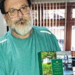 Homem de barba branca e óculos segura um exemplar do livro “Orquídeas Nativas de Florianópolis”. Ele veste uma camisa verde e está em um ambiente interno com janelas ao fundo, onde é possível ver persianas verticais. A capa do livro, voltada para a câmera, apresenta uma imagem de um cenário natural com uma casa simples e duas pessoas em um trapiche, além do título em destaque em letras brancas sobre um fundo verde.