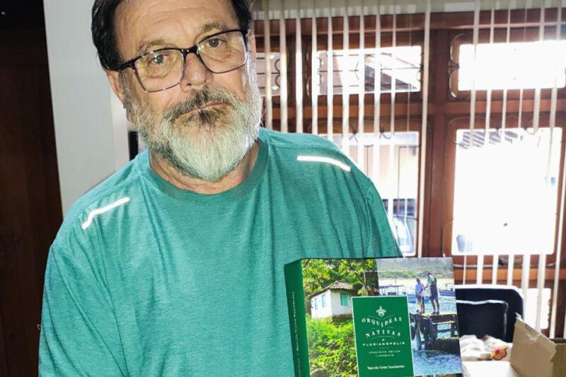 Homem de barba branca e óculos segura um exemplar do livro “Orquídeas Nativas de Florianópolis”. Ele veste uma camisa verde e está em um ambiente interno com janelas ao fundo, onde é possível ver persianas verticais. A capa do livro, voltada para a câmera, apresenta uma imagem de um cenário natural com uma casa simples e duas pessoas em um trapiche, além do título em destaque em letras brancas sobre um fundo verde.