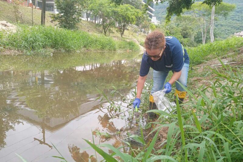 Fotografia de uma mulher coletando amostras de água em um rio raso, em uma área com vegetação verde nas margens. Ela usa luvas azuis, óculos escuros, calça jeans e botas amarelas, segurando um saco plástico transparente com uma das mãos e uma ferramenta de coleta com a outra. Ao fundo, há árvores, grama e construções parcialmente visíveis.