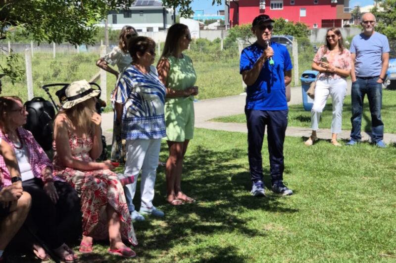 Fotografia em ambiente externo, com um grupo de pessoas em um gramado sob a sombra de uma árvore. Em primeiro plano, à esquerda, três mulheres estão sentadas; uma veste camisa xadrez rosa, outra usa vestido floral vermelho e chapéu claro, e a terceira está parcialmente encoberta. Ao centro, uma mulher de cabelos curtos veste blusa listrada azul e branca, ao lado de outra mulher de vestido verde-claro. À direita, um homem de boné preto, camiseta azul e calça escura segura um microfone enquanto fala. Ao fundo, há duas pessoas em pé, uma mulher de óculos com blusa estampada e calça branca, e um homem de barba com camiseta azul e jeans. O ambiente é iluminado pela luz do sol, com prédios e cercas ao fundo.