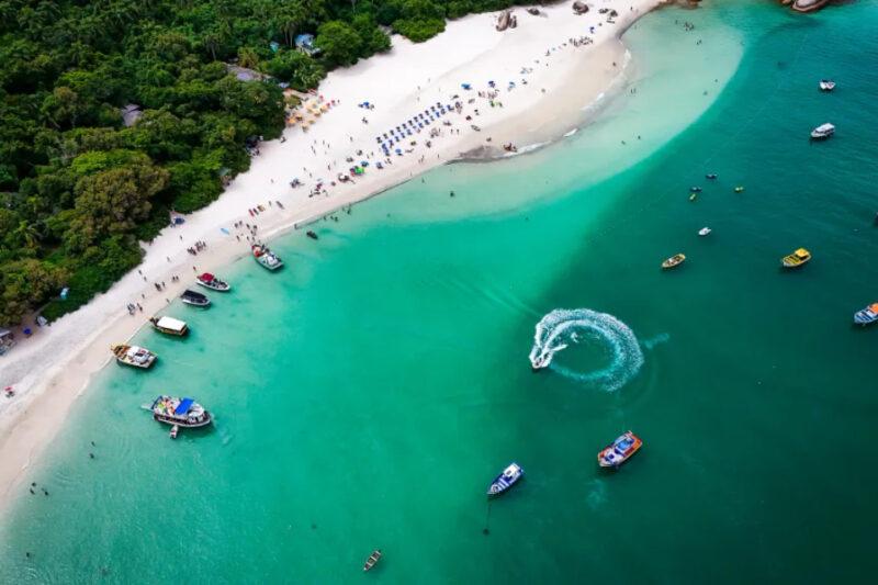 Fotografia aérea de uma praia de águas cristalinas em tons de verde esmeralda. Na areia clara, há várias pessoas espalhadas, algumas sob guarda-sóis coloridos alinhados. À esquerda, uma área de vegetação densa margeia a praia. No mar, várias embarcações pequenas, como barcos e lanchas, estão estacionadas ou em movimento, sendo que uma delas faz um giro que forma um rastro circular na água. O cenário é vibrante e repleto de atividade, transmitindo uma atmosfera tropical.