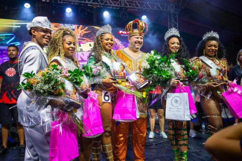 A imagem mostra seis pessoas em um palco, participando de um evento festivo, provavelmente relacionado ao Carnaval, todas vestindo trajes brilhantes e ornamentados típicos de desfiles ou concursos carnavalescos. À esquerda, um homem com um traje branco reluzente com detalhes em prata segura um buquê de flores; ao lado dele, duas mulheres com roupas douradas brilhantes, coroas e faixas seguram flores e sacolas de presentes. No centro, um homem mais velho veste um traje laranja brilhante com detalhes dourados, usa uma coroa dourada e segura flores e um troféu. À direita, outras duas mulheres usam trajes elaborados em tons de vermelho e dourado, com coroas e faixas, também segurando flores e sacolas de presentes. O fundo é composto por luzes de palco coloridas e uma tela com gráficos que remetem ao tema do evento, transmitindo um clima festivo e comemorativo.