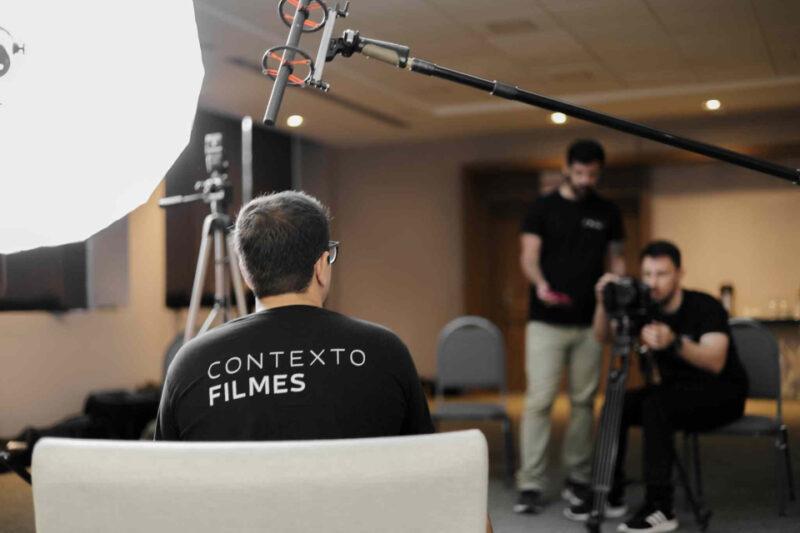 Fotografia dos bastidores de uma gravação em ambiente interno. Em primeiro plano, um homem visto de costas veste uma camiseta preta com a inscrição "CONTEXTO FILMES". À esquerda, há uma grande luz difusora, e no topo da imagem, um microfone boom está posicionado. Ao fundo, dois profissionais operam equipamentos de filmagem; um deles está manuseando uma câmera sobre tripé, enquanto o outro segura um dispositivo, possivelmente um controle remoto ou celular. O cenário sugere um ambiente profissional de produção audiovisual.