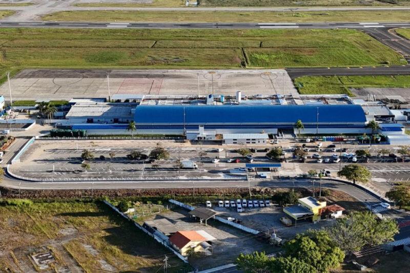 A imagem mostra uma vista aérea de um grande complexo hospitalar que anteriormente funcionava como um aeroporto, mantendo algumas características da sua antiga função, como extensas áreas pavimentadas e pistas desativadas ao fundo. No centro, destaca-se um galpão alongado com telhado azul, que parece ser o principal edifício do multihospital. Em frente a ele, há um amplo estacionamento com várias vagas disponíveis, algumas árvores e vias de acesso com veículos estacionados e em circulação. Atrás do edifício principal, ainda são visíveis áreas pavimentadas com marcações circulares, que podem ter sido usadas para operações aeroportuárias. Mais ao fundo, grandes áreas gramadas cercam as antigas pistas, agora sem uso para aviação. No canto inferior da imagem, há pequenas construções, incluindo uma casa com telhado vermelho e edificações menores, possivelmente de apoio ao hospital, além de um estacionamento menor com vários veículos brancos. O local foi adaptado para sua nova função hospitalar, preservando parte da infraestrutura original do aeroporto.