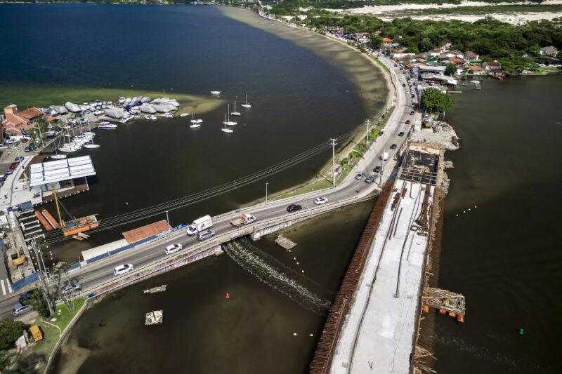 Fotografia aérea de uma ponte sobre um corpo d'água, com uma nova estrutura em construção ao lado da ponte existente. A via atual tem tráfego de carros e um caminhão, enquanto a ponte em construção exibe vigas de concreto e armações metálicas. À esquerda, há uma marina com barcos ancorados e cobertos, além de uma pequena estrutura com telhado vermelho. Do outro lado da ponte, uma estrada sinuosa acompanha a margem da água, passando por uma área arborizada e um pequeno bairro com casas. Ao fundo, dunas de areia contrastam com a vegetação.