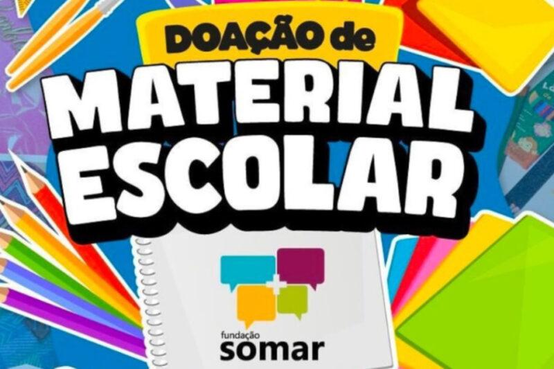 Imagem publicitária colorida promovendo a "Doação de Material Escolar". No centro, há a frase "Doação de Material Escolar" em letras grandes e brancas com bordas pretas, destacada por um fundo amarelo e preto. Abaixo, um caderno espiral com o logo da Fundação Somar, que inclui quatro balões de diálogo em diferentes cores (azul, verde, amarelo e roxo). Ao redor, há diversos itens escolares ilustrados, como lápis coloridos, borracha amarela, papel colorido e apontadores. O fundo é azul com padrões geométricos.