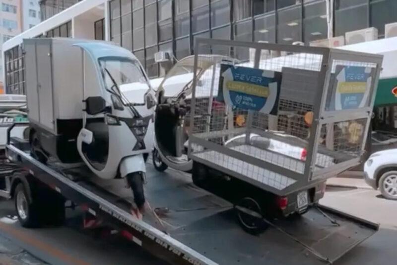 Fotografia de dois triciclos de carga elétricos sobre um caminhão plataforma em uma rua movimentada com prédios comerciais ao fundo. O triciclo à esquerda possui uma cabine fechada com compartimento de carga, enquanto o da direita tem uma carroceria aberta com estrutura metálica e placas que indicam "FEVER" e "Coleta Seletiva", sugerindo uso para reciclagem. O caminhão parece estar realizando o transporte ou remoção dos veículos.