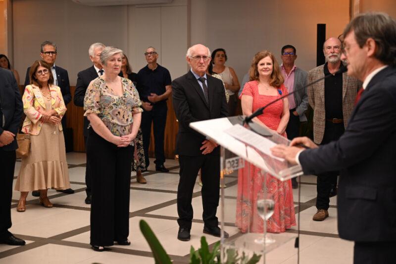 Homem de terno discursa em púlpito, enquanto grupo de pessoas o observa em ambiente formal.