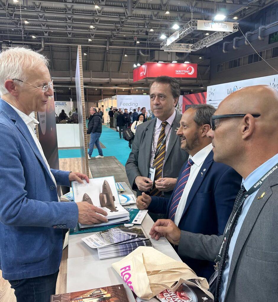 Homem de blazer azul mostra um livro aberto para três homens de terno em um evento profissional.