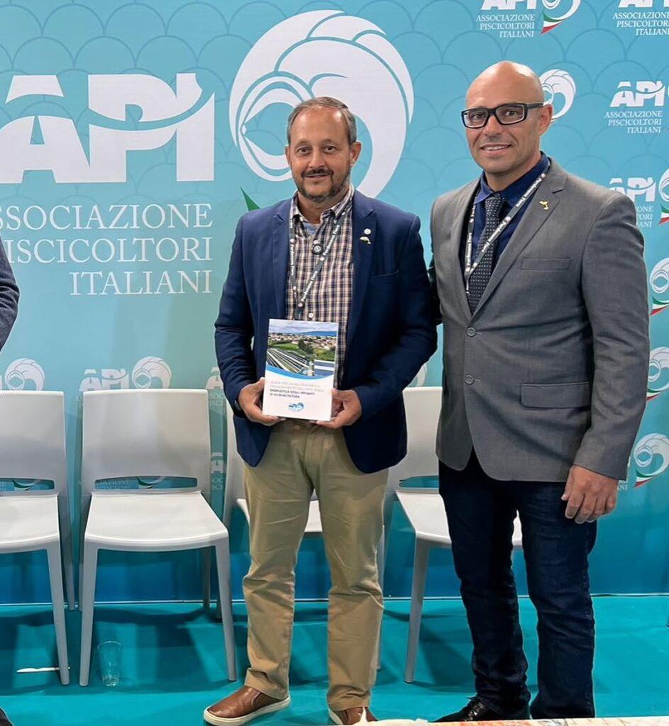 Dois homens de terno posam ao lado de um terceiro segurando um livro. Fundo com painel da Associação Piscicultores Italianos.