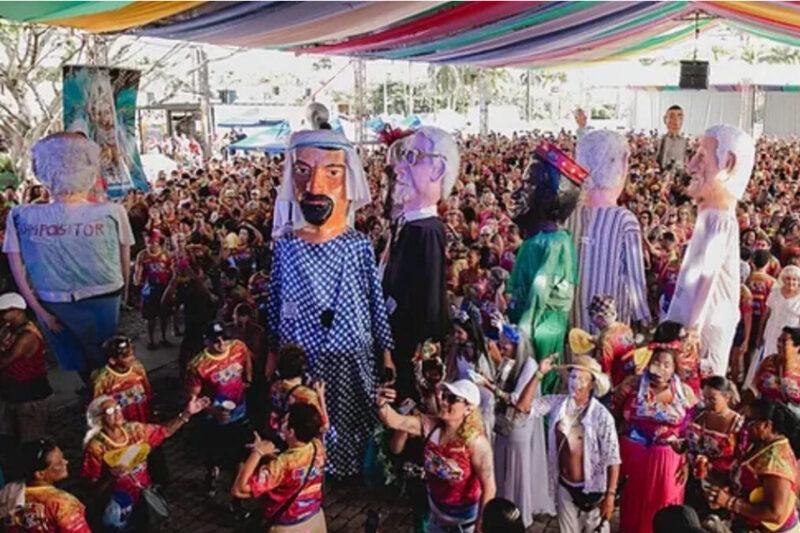 Desfile de bonecos gigantes em meio a uma multidão festiva, sob um toldo colorido.