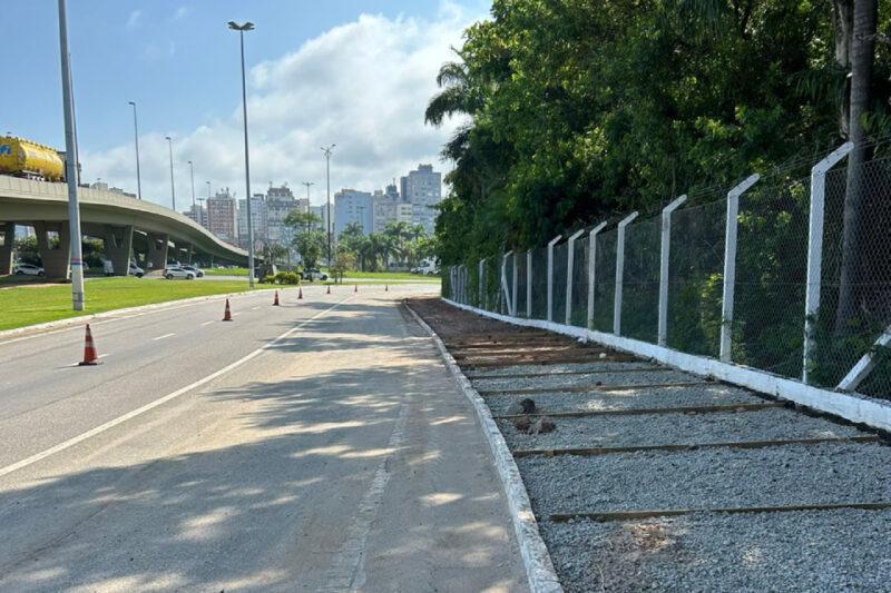 Uma rua parcialmente interditada com cones laranjas espaçados ao longo da faixa lateral. À direita, há uma calçada em construção, coberta com brita e delimitada por guias brancas desgastadas. Ao lado da calçada, uma cerca de arame com postes brancos separa a área arborizada da via. No fundo, vê-se um viaduto, prédios e céu azul com algumas nuvens.