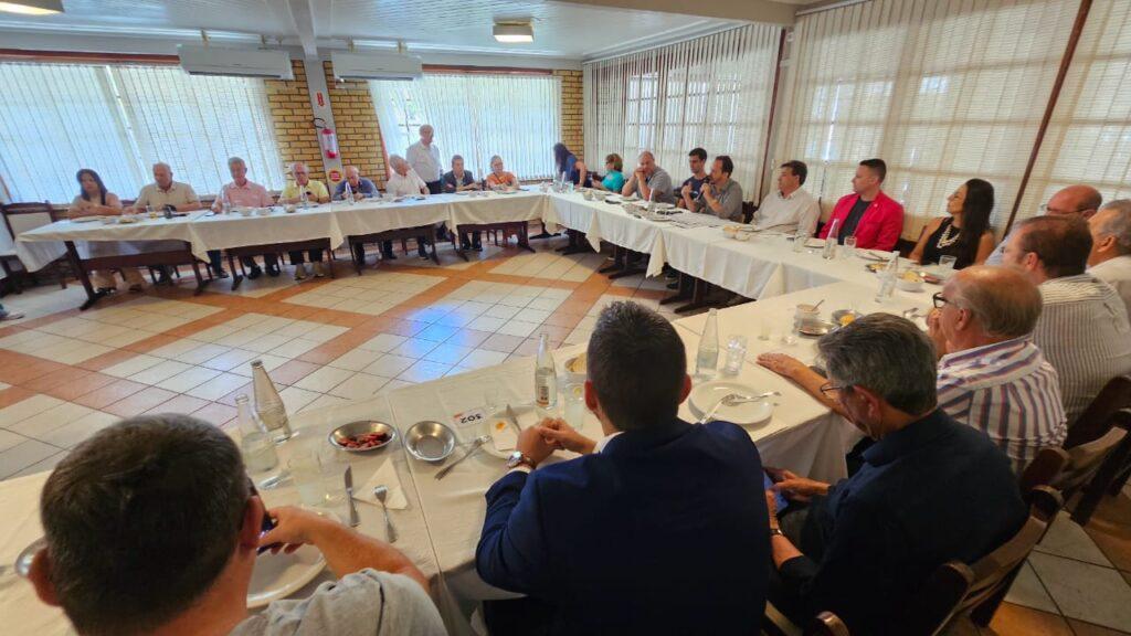 Vista da reunião com participantes atentos. Mesa com pratos, copos e garrafas. Fundo com janelas e cortinas. Pessoas sentadas em mesa posta com comida, enquanto outras conversam. Ambiente amplo com luz natural.