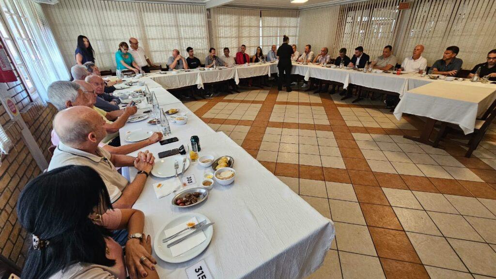 Pessoas sentadas em mesa posta com comida, enquanto outras conversam. Ambiente amplo com luz natural.