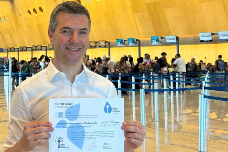 Homem sorridente segura um certificado em um aeroporto movimentado, próximo a balcões de check-in.