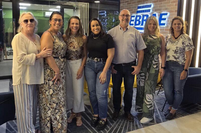 Sete pessoas posam sorridentes para a foto em um ambiente interno com decoração moderna. No fundo, há uma parede de tijolos com um letreiro iluminado escrito "SEBRAE". O grupo é composto por seis mulheres e um homem, vestidos com roupas casuais e sociais. Algumas mulheres usam vestidos estampados, enquanto outras estão de calça e blusa. O homem veste uma camisa clara e calça escura. O grupo está próximo, com alguns se abraçando ou tocando nos ombros uns dos outros.