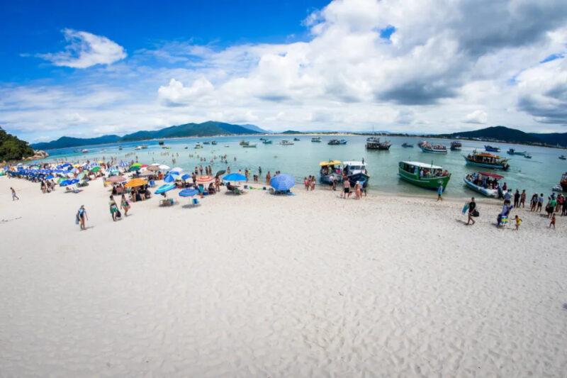 Praia movimentada com areia branca e mar calmo, onde várias pessoas aproveitam o dia ensolarado. Há guarda-sóis coloridos espalhados pela areia e grupos de banhistas dentro d'água. Barcos de diferentes tamanhos estão ancorados próximos à margem, alguns transportando turistas. Ao fundo, montanhas cobertas de vegetação cercam a paisagem, e o céu azul tem nuvens brancas dispersas.