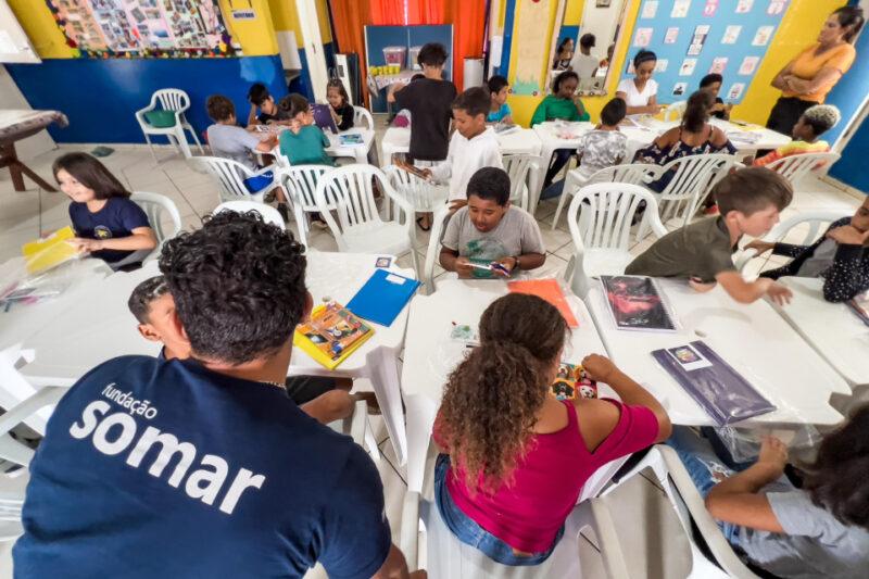 Crianças sentadas em mesas brancas participam de uma atividade educativa em uma sala colorida, decorada com murais e cartazes. Algumas estão manipulando materiais escolares, enquanto um homem de camisa azul com a inscrição "Fundação Somar" interage com um menino. Ao fundo, uma mulher de blusa laranja observa a cena.