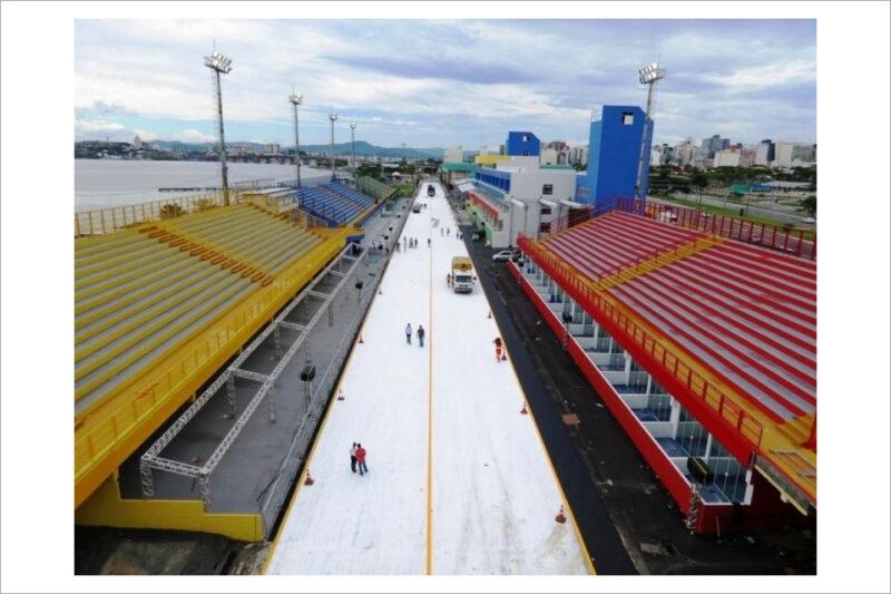 Vista aérea de um sambódromo com arquibancadas coloridas e poucas pessoas na pista em preparação.