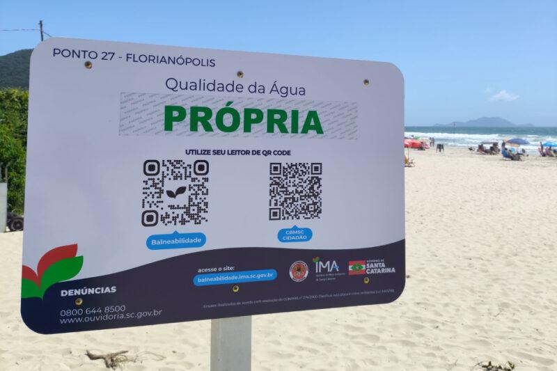 Placa informativa na praia em Florianópolis indica que a qualidade da água é "PRÓPRIA" para banho. A placa contém dois QR codes, informações sobre balneabilidade e contato para denúncias. Ao fundo, a praia tem areia clara, mar azul com ondas e algumas pessoas sob guarda-sóis coloridos.