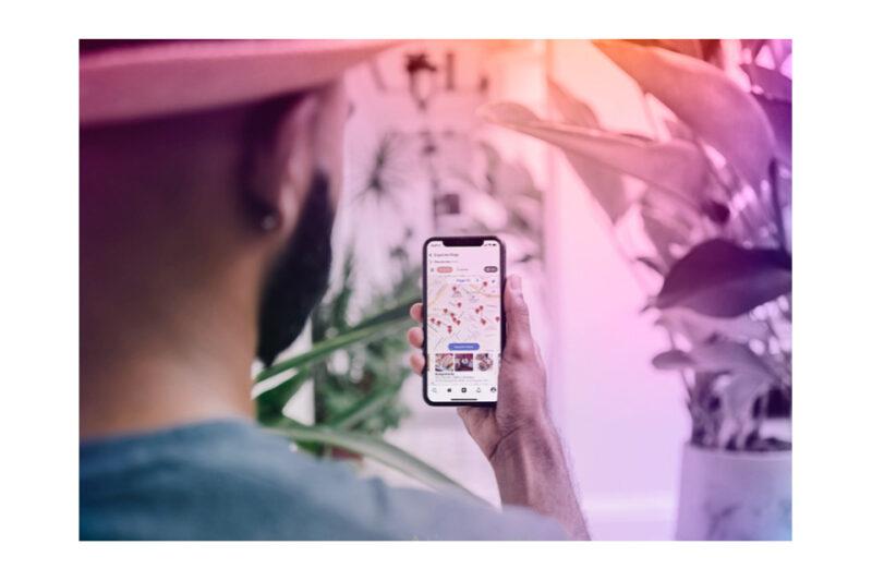 Homem de chapéu visto por trás segura um smartphone com um mapa aberto na tela. O ambiente ao redor tem plantas e iluminação em tons rosados.