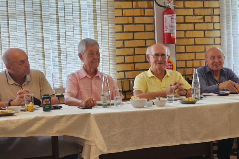 Quatro homens idosos sentados à mesa, sorrindo, com comida e bebida. Fundo com tijolos e extintor.