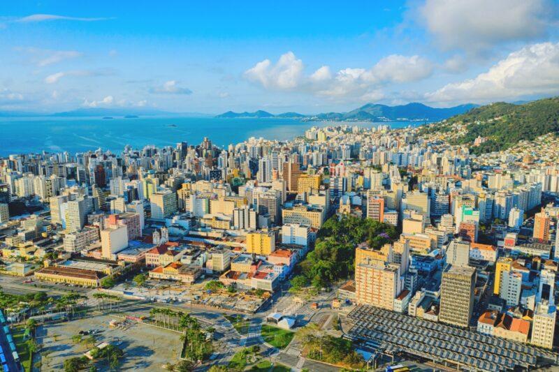 Vista aérea de Florianópolis, Santa Catarina, mostrando a área central da cidade com prédios altos, ruas movimentadas e o Terminal de Integração do Centro. Ao fundo, o mar azul e montanhas verdes compõem a paisagem. O céu está claro, com algumas nuvens brancas.