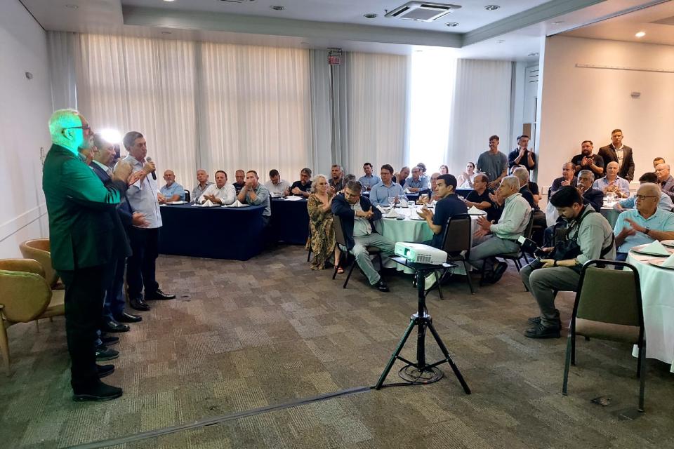 Homens falam ao microfone diante de uma plateia atenta em um evento corporativo, com um projetor à frente.