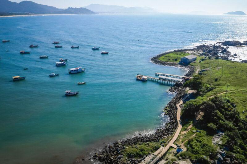 Vista aérea de uma enseada com mar azul, barcos ancorados, um píer de madeira e uma área verde com trilhas.