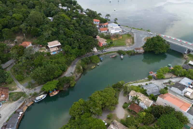 Vista aérea de um canal cercado por vegetação, casas e barcos, próximo a uma estrada e uma ponte.