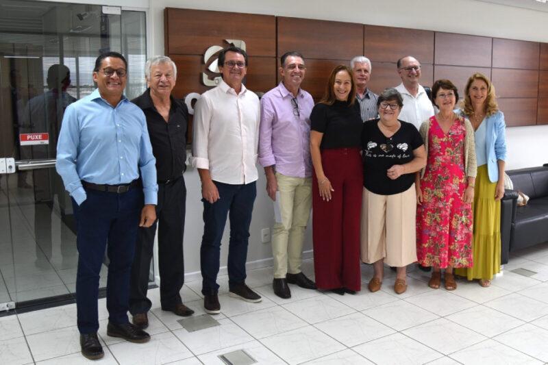 Grupo de dez pessoas sorri em um saguão corporativo, posando diante de um painel de madeira com logotipo.