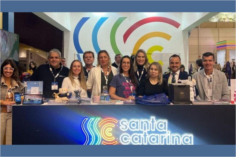 Grupo de pessoas sorrindo atrás de um balcão iluminado com o logo "Santa Catarina" em um evento.