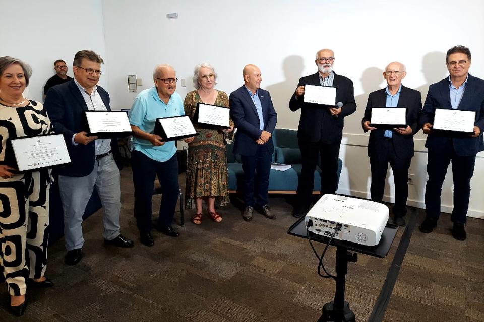 Grupo de pessoas posa segurando certificados em evento de premiação, com um projetor à frente e parede branca ao fundo.