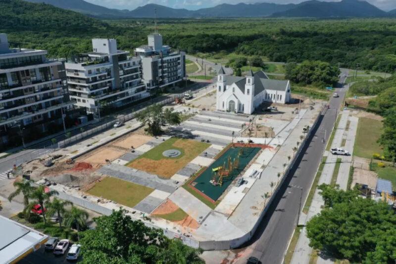 Vista aérea de uma praça em construção com igreja branca, playground e prédios modernos ao fundo.