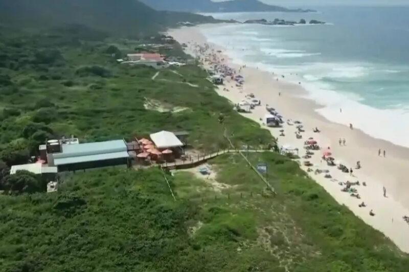 Vista aérea de uma praia movimentada com areia clara, mar azul e vegetação densa ao redor de quiosques.