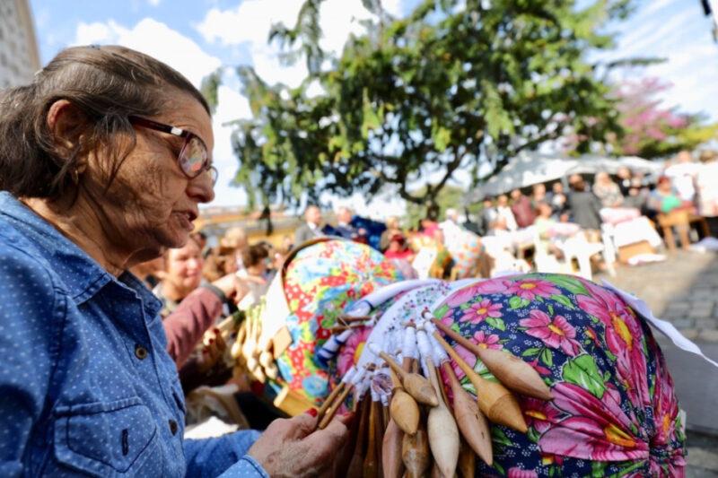 Mulher idosa faz renda de bilros ao ar livre, rodeada por outras rendeiras e espectadores.