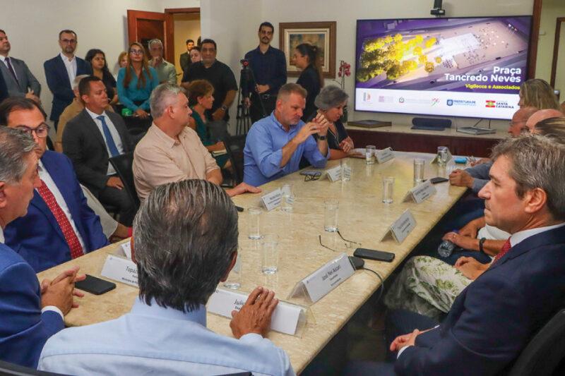 Reunião com autoridades em torno de mesa. Tela ao fundo exibe projeto da Praça Tancredo Neves.