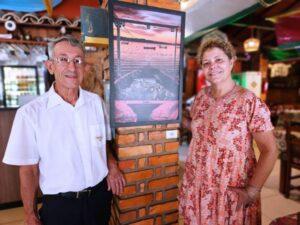 Homem e mulher sorriem ao lado de uma foto de barco com peixes ao pôr do sol, fixada na parede.