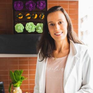 Mulher sorridente de blazer branco em frente a parede com flores decorativas coloridas.