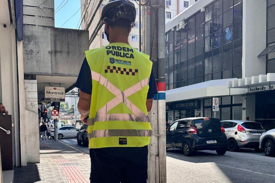 Agente de Ordem Pública com colete refletivo observa o trânsito em rua movimentada de Florianópolis.