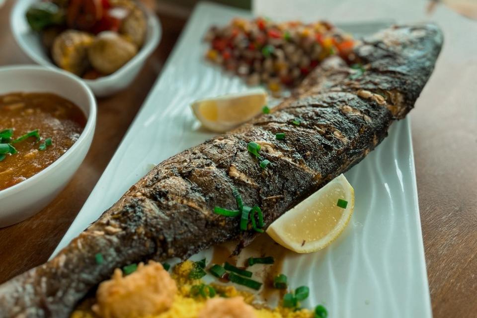 Peixe grelhado inteiro com limões, farofa, vinagrete e acompanhamentos em tigelas ao fundo.