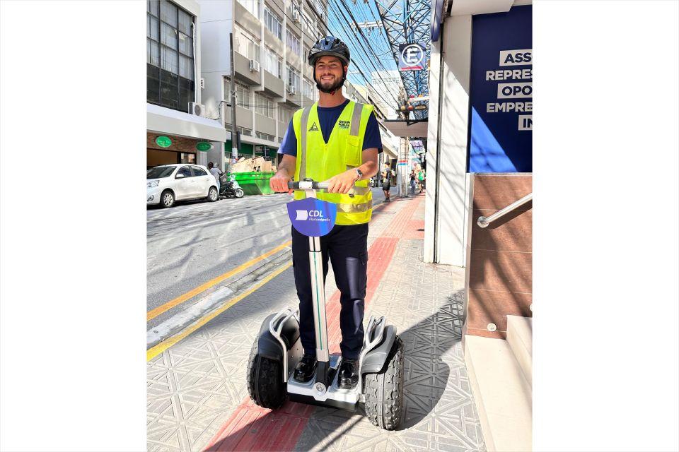 Homem sorridente com colete amarelo e capacete anda de patinete elétrico na calçada de rua urbana.