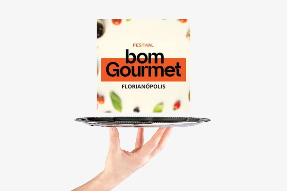 Cartaz do Festival Bom Gourmet Florianópolis sobre uma bandeja, com fundo claro e frutas desfocadas.