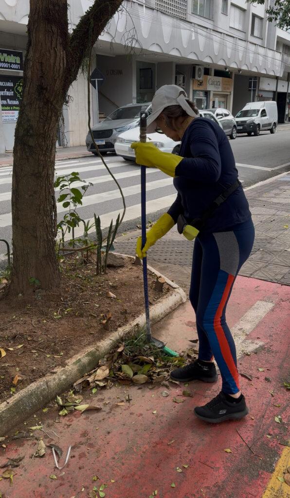 Mulher com luvas e boné varre folhas secas próximo a faixa de pedestre.