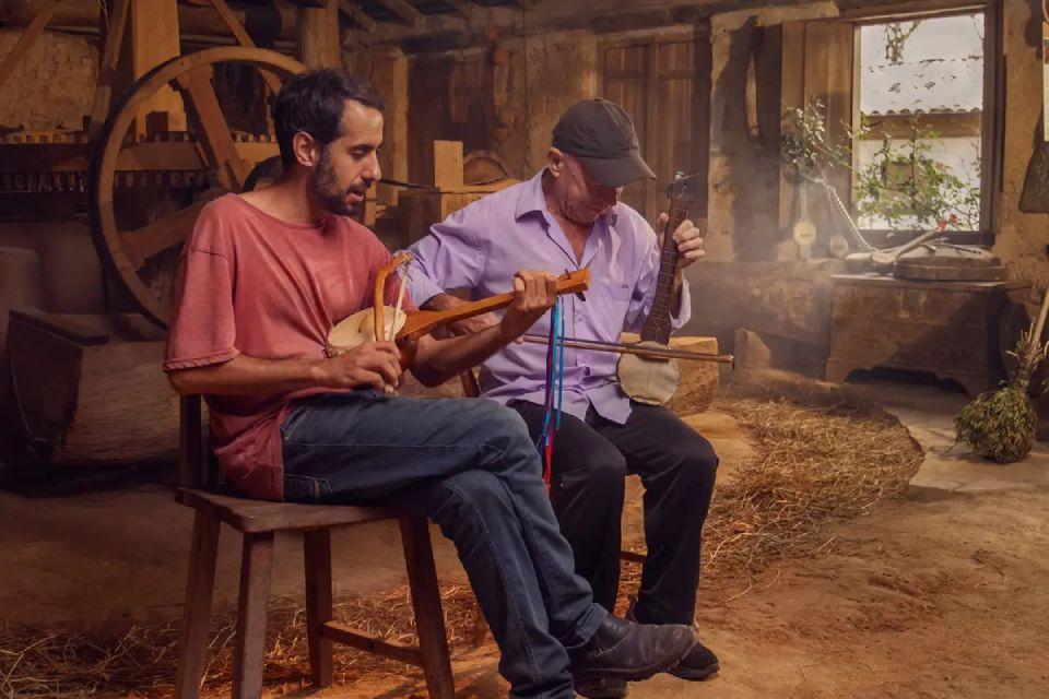 Fotografia de dois homens sentados em um ambiente rústico, tocando instrumentos de corda artesanais.