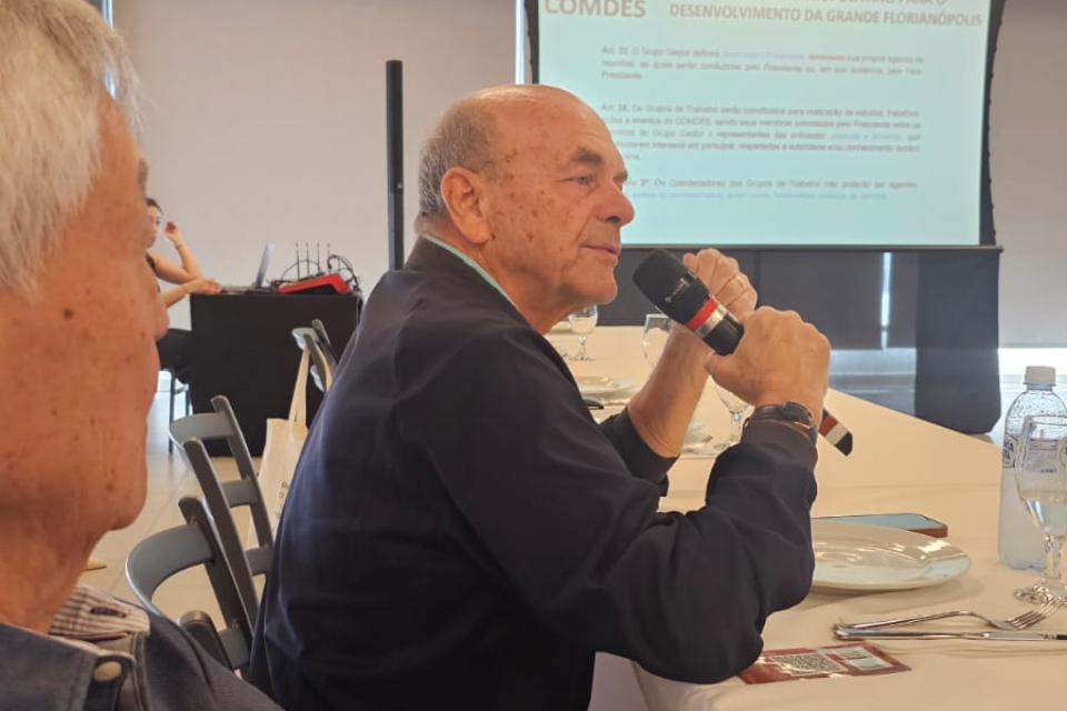 Homem calvo segura microfone e fala em reunião, com telão ao fundo exibindo texto sobre Florianópolis.